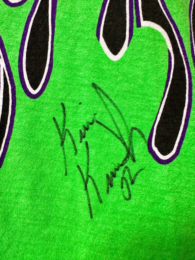 May include: Close-up of a vibrant green t-shirt with a flame design in black, purple, and white. The shirt has a handwritten signature in black ink.