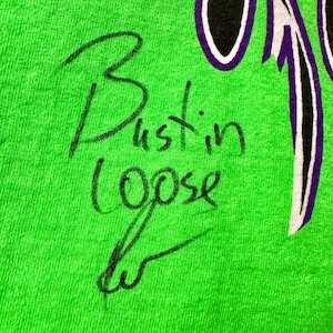 May include: Close-up of a vibrant green fabric featuring the handwritten phrase "Bustin Loose" in black ink. A purple and white design is visible in the upper right corner. The fabric has a slightly textured appearance.