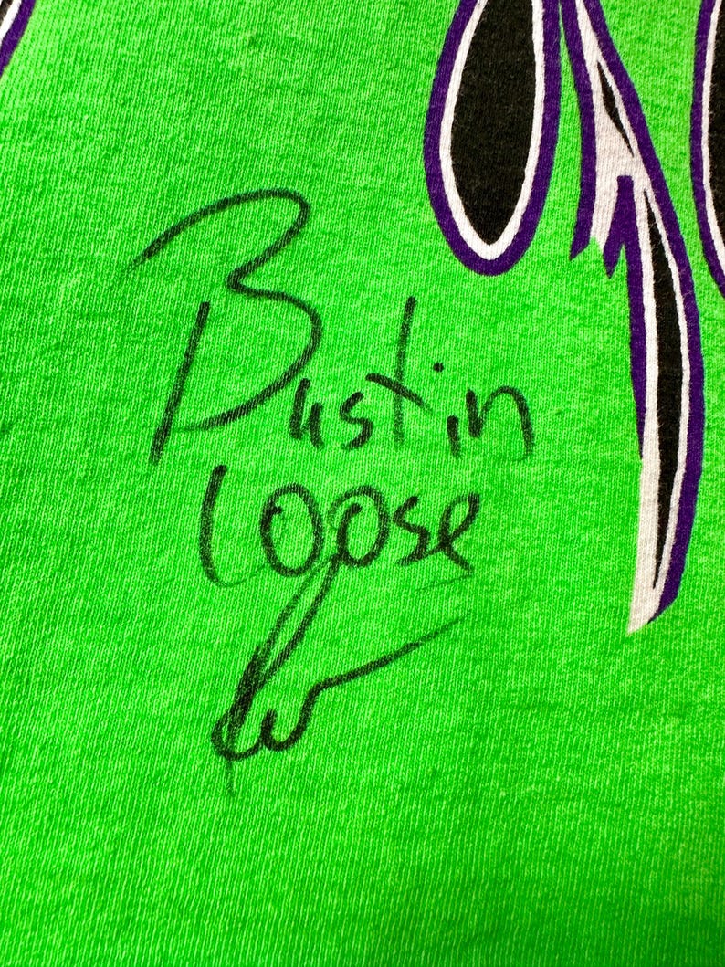 May include: Close-up of a vibrant green fabric featuring the handwritten phrase "Bustin Loose" in black ink. A purple and white design is visible in the upper right corner. The fabric has a slightly textured appearance.