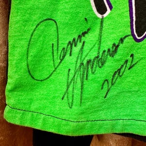 May include: Close-up of a neon green t-shirt with a black and white design. The shirt features a handwritten signature in black ink, along with the number "2002". The bottom of the shirt is black.