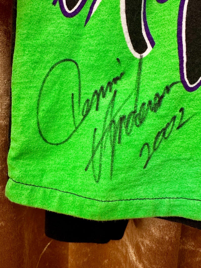 May include: Close-up of a neon green t-shirt with a black and white design. The shirt features a handwritten signature in black ink, along with the number "2002". The bottom of the shirt is black.