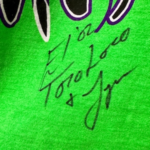 May include: Close-up of a bright green t-shirt with a black and purple design at the top. The shirt has a handwritten signature in black ink. The signature includes the name "El Loco Toro Jr."