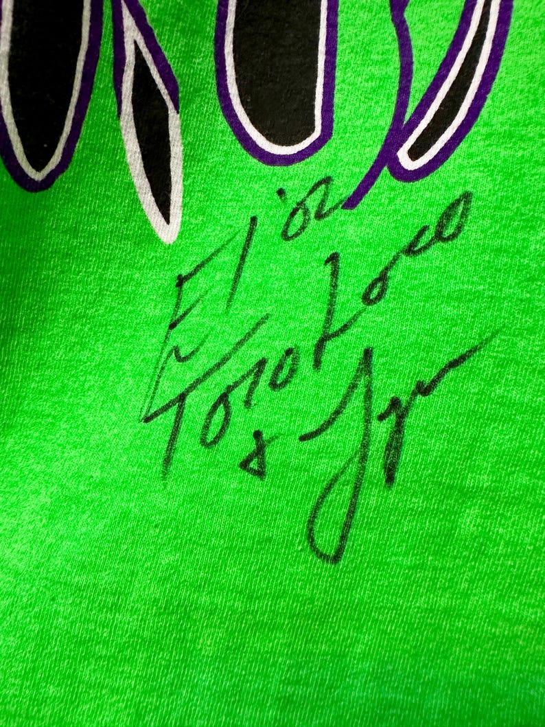 May include: Close-up of a bright green t-shirt with a black and purple design at the top. The shirt has a handwritten signature in black ink. The signature includes the name "El Loco Toro Jr."