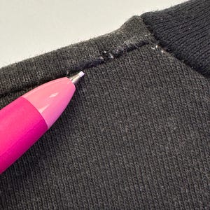 May include: Close-up of dark grey knit fabric with a pink and purple pen pointing at the seam. The fabric has a ribbed texture, and the seam shows white stitching.