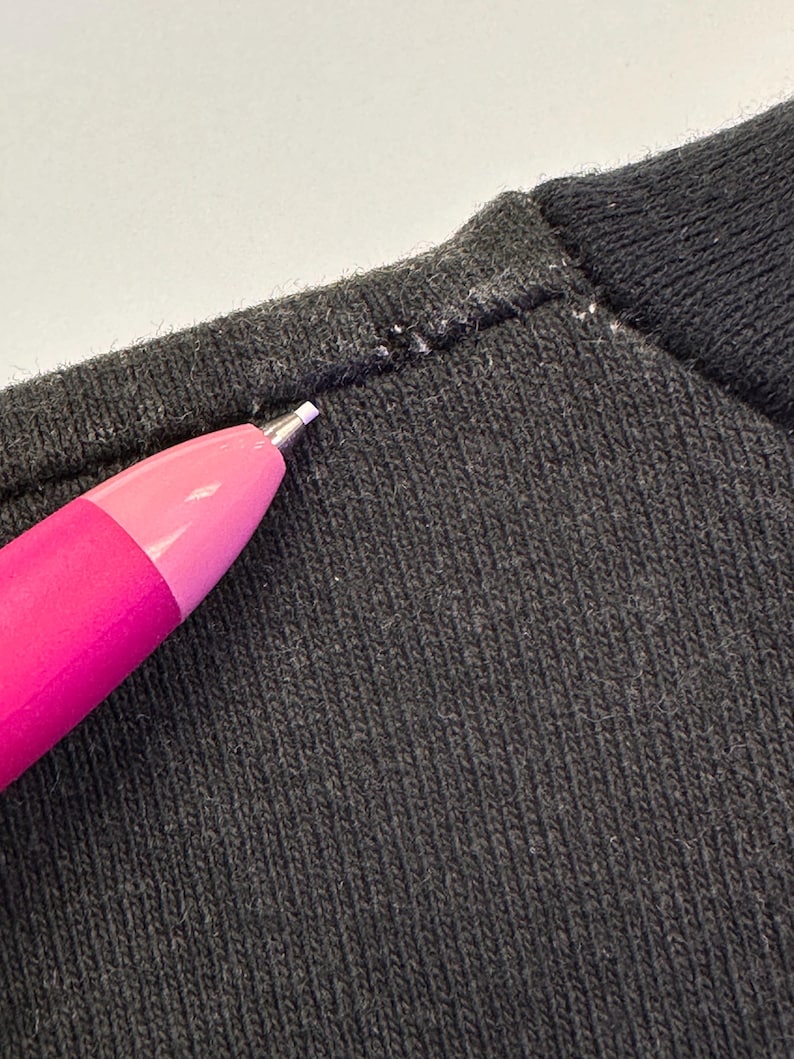 May include: Close-up of dark grey knit fabric with a pink and purple pen pointing at the seam. The fabric has a ribbed texture, and the seam shows white stitching.