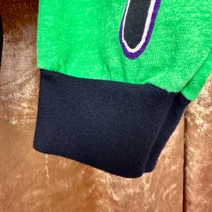 May include: Close-up of a green long-sleeve shirt sleeve with a black cuff. The shirt features a black design with a white and purple outline. The fabric has a textured appearance.