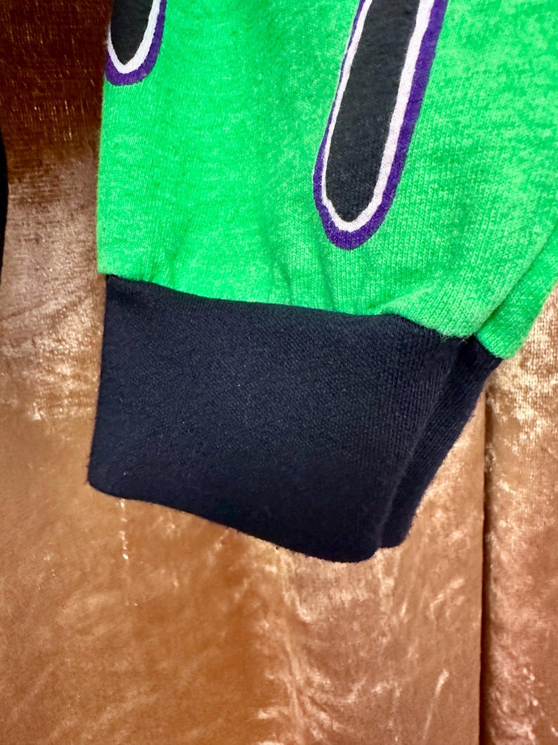May include: Close-up of a green long-sleeve shirt sleeve with a black cuff. The shirt features a black design with a white and purple outline. The fabric has a textured appearance.