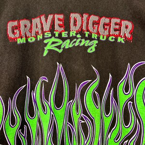 May include: Dark grey sweatshirt with the text "GRAVE DIGGER MONSTER TRUCK Racing" in red, grey, and green. The bottom of the sweatshirt has a flame design in green, purple, and white.
