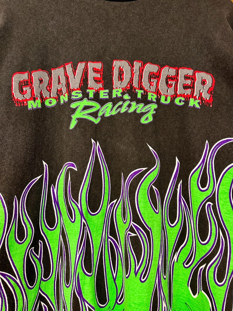 May include: Dark grey sweatshirt with the text "GRAVE DIGGER MONSTER TRUCK Racing" in red, grey, and green. The bottom of the sweatshirt has a flame design in green, purple, and white.