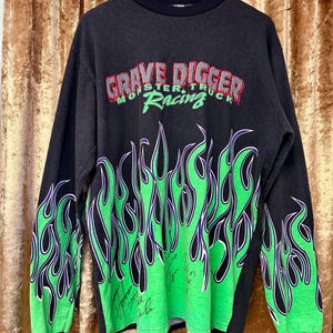 May include: Black long-sleeved top with the text "GRAVE DIGGER MONSTER TRUCK RACING" in red and green. It has green and purple flame graphics on the sleeves and lower front.
