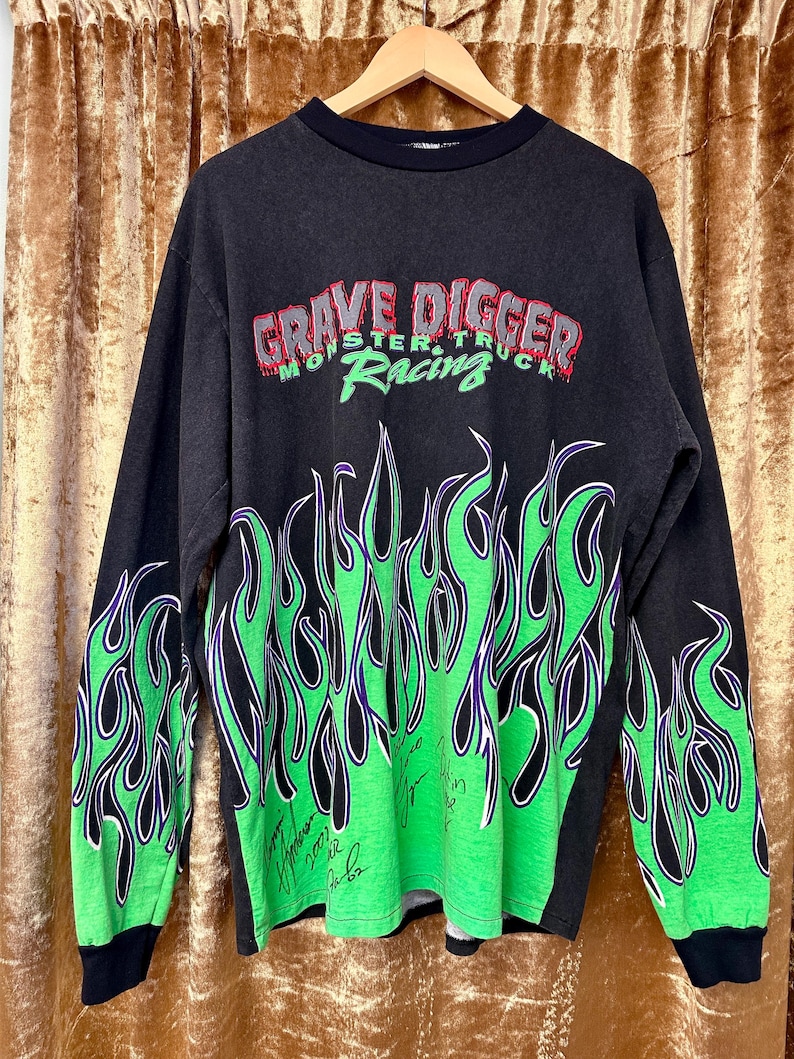 May include: Black long-sleeved top with the text "GRAVE DIGGER MONSTER TRUCK RACING" in red and green. It has green and purple flame graphics on the sleeves and lower front.