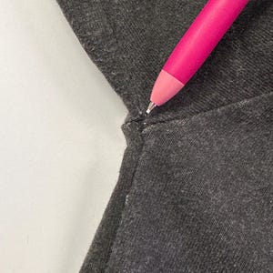 May include: A close-up shot of dark grey fabric with a seam. A bright pink pen, decorated with a floral pattern, is angled towards the fabric. The pen's tip is visible, emphasizing the texture and detail of the material.