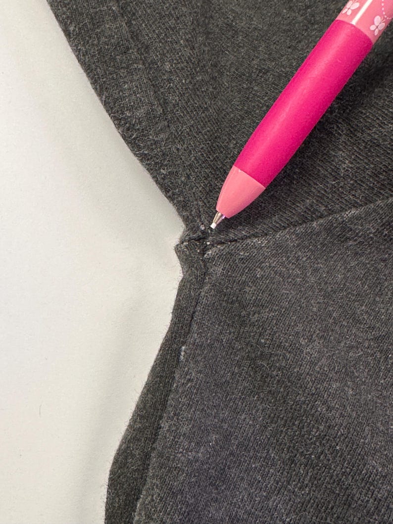 May include: A close-up shot of dark grey fabric with a seam. A bright pink pen, decorated with a floral pattern, is angled towards the fabric. The pen's tip is visible, emphasizing the texture and detail of the material.