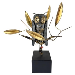 May include: A metal sculpture of an owl perched on a branch with gold leaves. The owl is made of various metal pieces and has a black and silver finish. The sculpture is mounted on a black base.