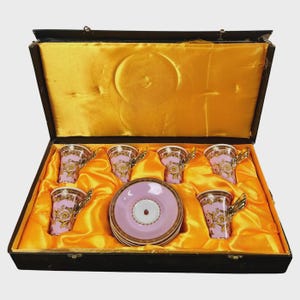 May include: A set of six pink teacups and saucers with gold accents, presented in a black box with a gold satin lining. The teacups feature a decorative gold design and gold handles. The saucers are pink with a gold trim.