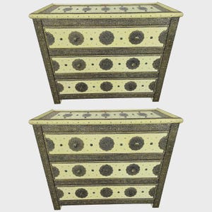 May include: Two cream-colored chests of drawers with intricate metalwork and decorative circular accents. The chests have four drawers each and are set against a neutral background. The metalwork is a dark, antique gold color.