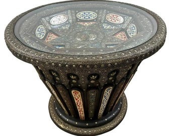 May include: A round, ornate coffee table with a glass top. The table is made of wood and metal, and features intricate carvings and inlaid designs. The table is decorated with colorful geometric patterns and has a dark brown finish.