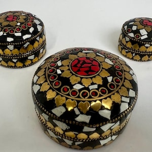 May include: Three decorative round boxes with lids. The boxes are black with gold, white, and red mosaic-style embellishments. The largest box is in the foreground, with two smaller boxes behind it.