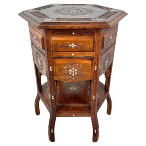 May include: A hexagonal wooden table with intricate carvings and inlaid mother-of-pearl accents. The table has three drawers and a lower shelf. The table is made of dark wood and has a rich, warm finish.
