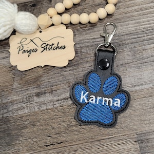 May include: Blue paw print keychain with the name "Karma" embroidered in white on a black leather background. The keychain has a silver metal clasp.