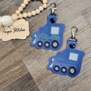May include: Two blue train-shaped keychains with silver clasps. The keychain on the left has the name Hayden embroidered in green on the front.