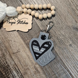 May include: A gray heart-shaped keychain with black and silver stripes and the number 8292 embroidered on it. The keychain has a silver metal ring and a black button.