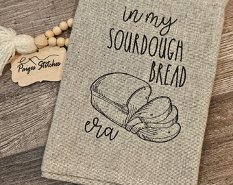 May include: A beige kitchen towel with black embroidery that reads "In my sourdough bread era" and features a drawing of a loaf of bread and slices of bread.
