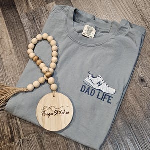 Puede incluir: Camiseta azul claro con las palabras "DAD LIFE" y un gr&aacute;fico de zapatillas deportivas. Una guirnalda de cuentas de madera y una etiqueta redonda de madera con el texto "Paiges Stitches" tambi&eacute;n est&aacute;n en la imagen.