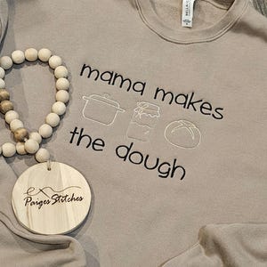 May include: A tan sweatshirt with the text "mama makes the dough" embroidered in black. The text is accompanied by three small embroidered illustrations: a pot, a jar, and a bun. A small wooden tag with the text "Ex Paiges Stitches" is attached to the sweatshirt.