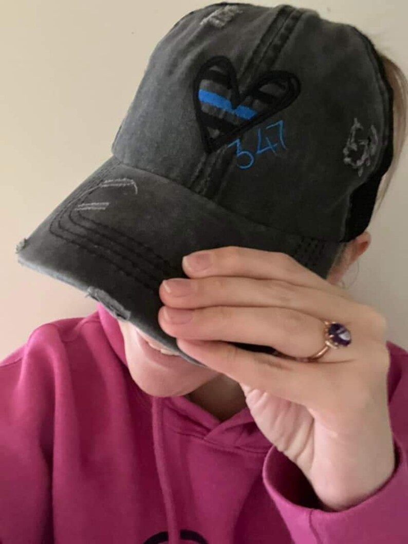 May include: A black distressed baseball cap with a blue and black heart design and the number 347 embroidered on the front. The cap has a mesh back.