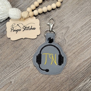 May include: Gray leather keychain with a black headphone design and the initials "TN" embroidered in yellow thread.