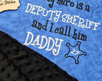May include: A blue and black baby blanket with the embroidered text "My hero is a DEPUTY SHERIFF and I call him DADDY" with a white star and the number 27.