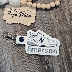 May include: White and blue embroidered shoe keychain with the name "Emerson" on a white background.