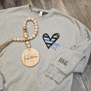 May include: Gray sweatshirt with a black and blue heart with the number 576 embroidered on it. The text "DUHIG" is embroidered on the sleeve. A wooden tag with the text "Paiges Stitches" is on the sweatshirt.