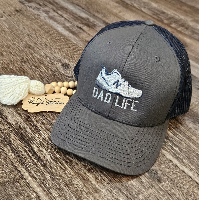 Puede incluir: Gorra de camionero gris con parte trasera de malla azul marino. La gorra presenta una imagen bordada de una zapatilla deportiva y las palabras "DAD LIFE" en blanco. La gorra est&aacute; sobre una superficie de madera.