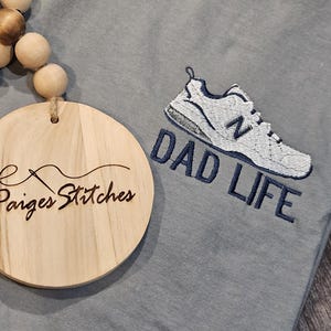 Puede incluir: Camiseta azul claro con un dise&ntilde;o bordado de zapatilla deportiva en blanco y azul marino y las palabras "DAD LIFE". Un adorno redondo de madera con el texto "Paiges Stitches" es visible.