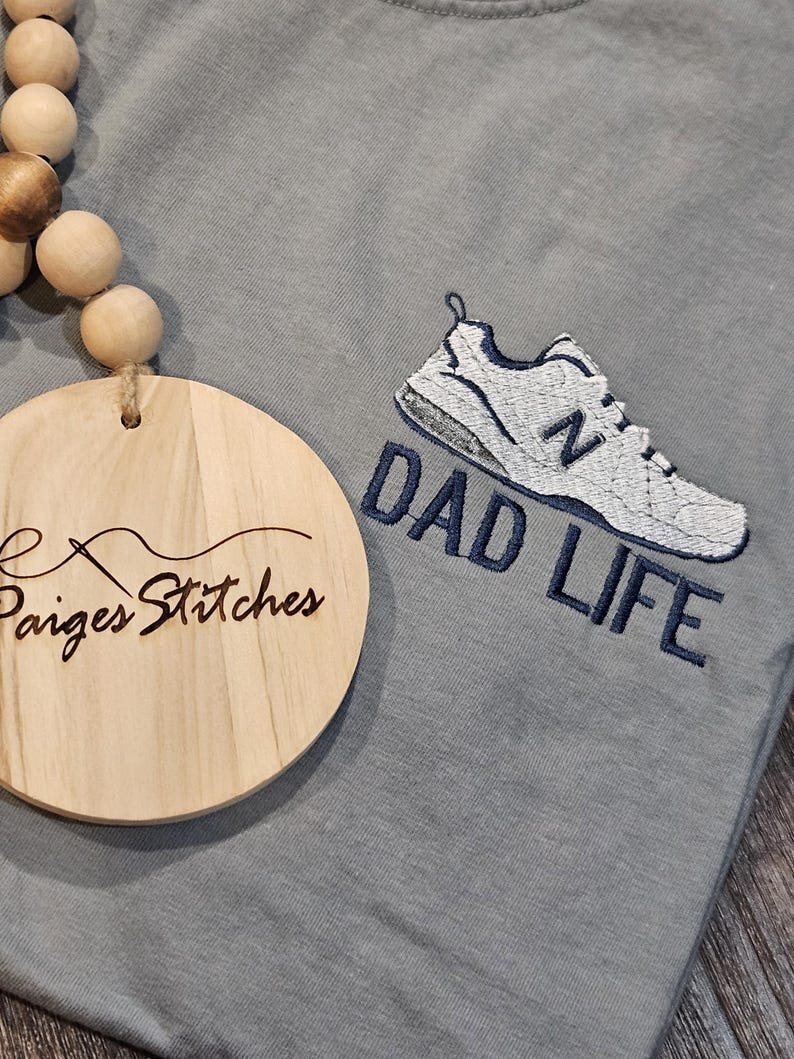 Puede incluir: Camiseta azul claro con un dise&ntilde;o bordado de zapatilla deportiva en blanco y azul marino y las palabras "DAD LIFE". Un adorno redondo de madera con el texto "Paiges Stitches" es visible.