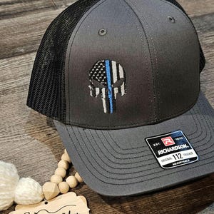 May include: A gray and black trucker hat with a blue line American flag skull design. The hat has an adjustable strap and a Richardson logo with the text "EST. 1970 ORIGINAL 112 TRUCKER ADJUSTABLE".
