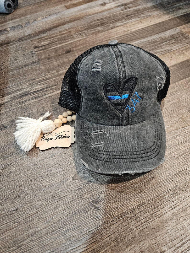 May include: A black and gray distressed trucker hat with a blue line heart and the number 347 embroidered on the front. The hat has a mesh back.
