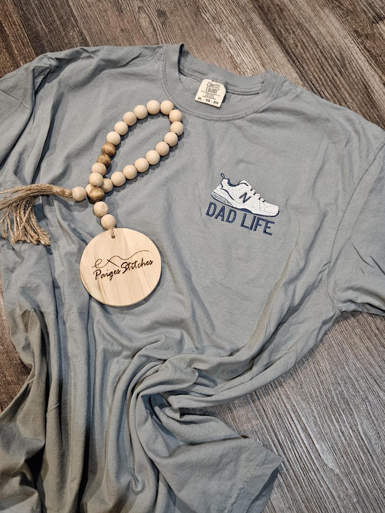 Puede incluir: Una camiseta gris Comfort Colors con el texto "DAD LIFE" y un gr&aacute;fico de zapato. Una guirnalda de cuentas de madera y un adorno de madera con el texto "Paiges Stitches" tambi&eacute;n est&aacute;n en la imagen.