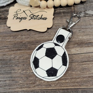 May include: A white and black soccer ball keychain with a silver clasp. The keychain is round and features a classic soccer ball design. The text "Paiges Stitches" is on a wooden tag.