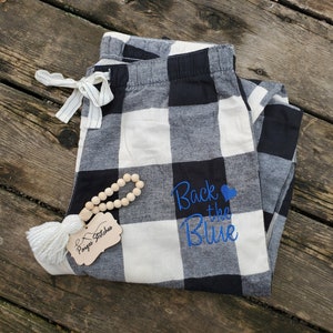 May include: Black and white buffalo plaid pajama pants with blue embroidery that says "Back the Blue" with a blue heart. The pants are folded and lying on a wooden surface.
