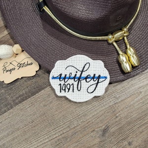 May include: A white embroidered patch with the word "wifey" in cursive, a blue line, and the number "1491". The patch is on a wooden surface next to a brown hat with a gold band and tassels. The patch is a handmade item.