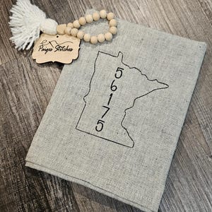 May include: A light gray fabric item with a black outline of the state of Minnesota and the numbers 56175. A wooden tag with the text "Paiges Stitches" hangs from a beaded garland and a white tassel.