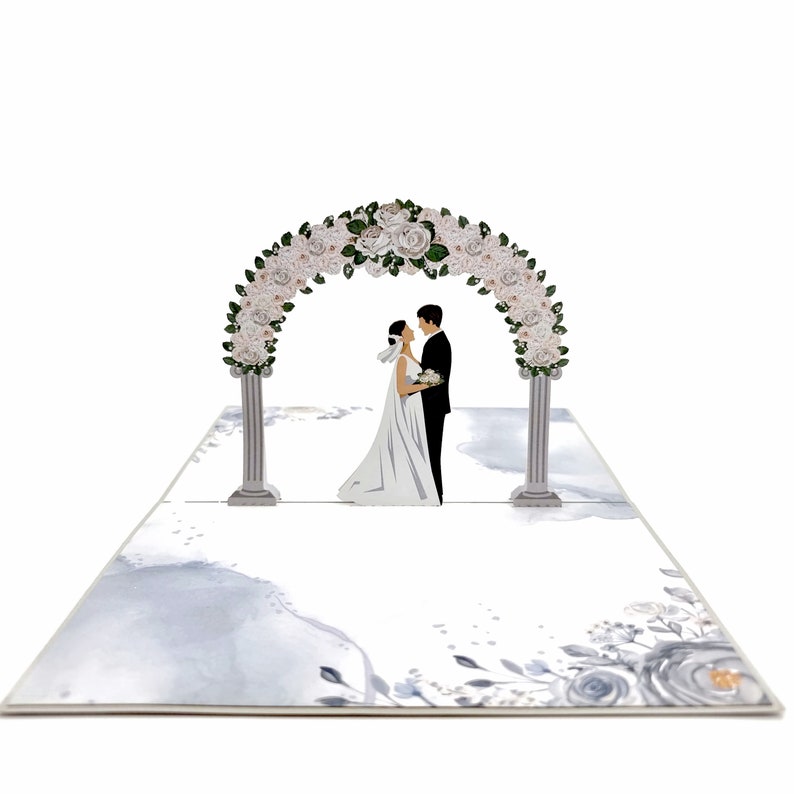 Op de afbeelding: Een 3D pop-up kaart die een huwelijksceremonie voorstelt met een stel dat onder een bloemenboog staat. De boog is gemaakt van witte bloemen en groen, en het stel is gekleed in formele kleding. De achtergrond is een aquarelontwerp met een lichtblauw en grijs kleurenschema.
