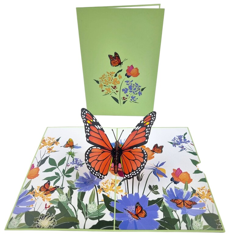 Peut inclure: Une carte pop-up 3D repr&eacute;sentant un papillon monarque aux ailes orange et noires, entour&eacute; de fleurs color&eacute;es. La carte est ferm&eacute;e et montre un fond vert avec un papillon plus petit.