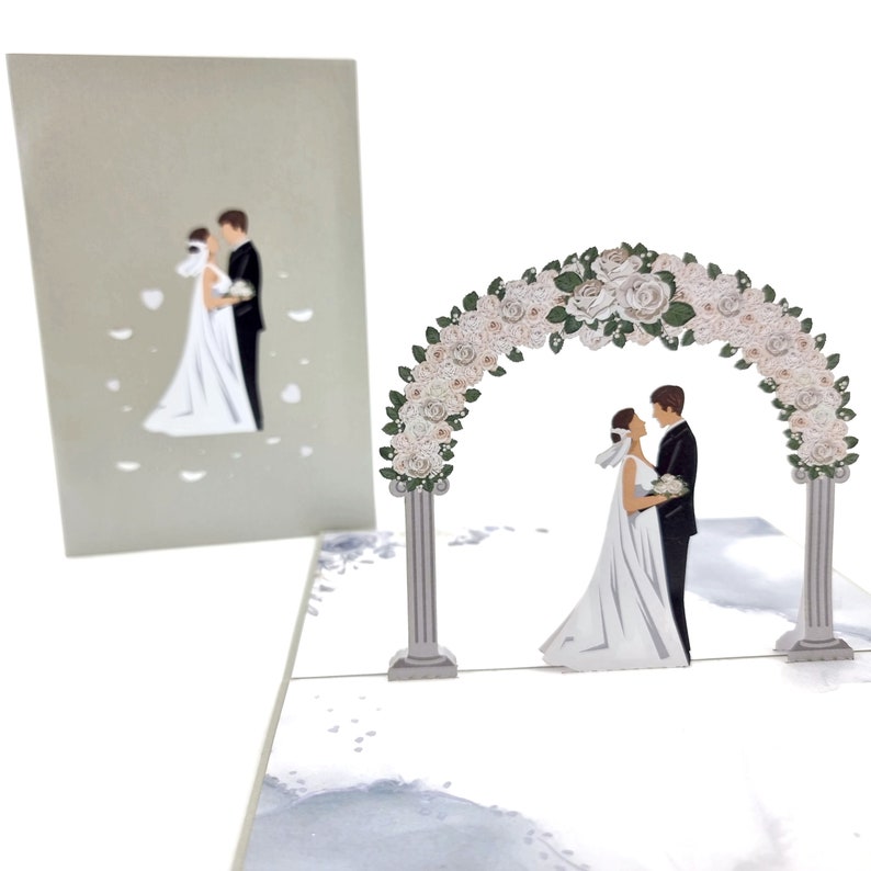 Op de afbeelding: Een 3D pop-up kaart die een huwelijksceremonie voorstelt. De kaart heeft een witte bloemenboog met een stel dat eronder staat. Het stel is gekleed in formele kleding, de bruid in een witte jurk en de bruidegom in een zwart pak. De kaart is perfect voor een huwelijks- of verlovingviering.