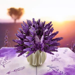 May include: A display of vivid purple lavender flowers in a cream-coloured, petal-shaped vase. The arrangement is on a table with a lavender-themed tablecloth. The background shows a soft, blurred landscape with a tree and a sunset.