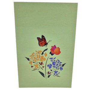 Peut inclure: Une carte 3D pop-up repr&eacute;sentant un papillon en vol au-dessus d'un bouquet de fleurs. Le papillon est orange et noir, et les fleurs sont violettes, orange et rouges.