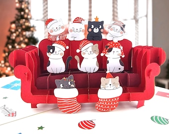 May include: A three-dimensional red paper sofa pop-up card featuring nine illustrated cats wearing festive holiday hats. Two cats are tucked into red and white striped stockings at the base, while seven others sit on the sofa cushions.